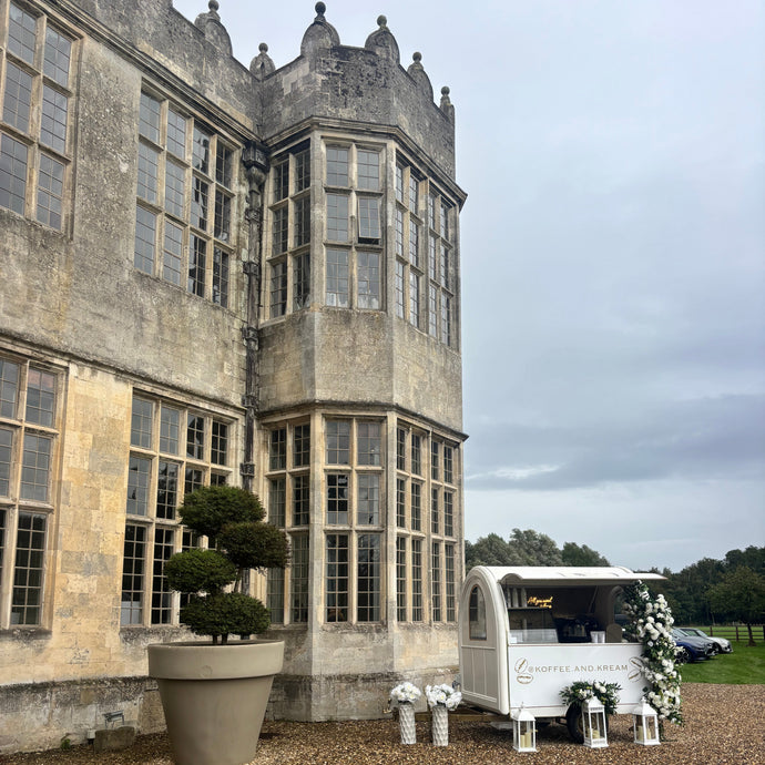 A white vintage-style wedding cart decorated with white flowers, set on a gravel courtyard in front of a large historic stone mansion Howsham hall  under an overcast sky.